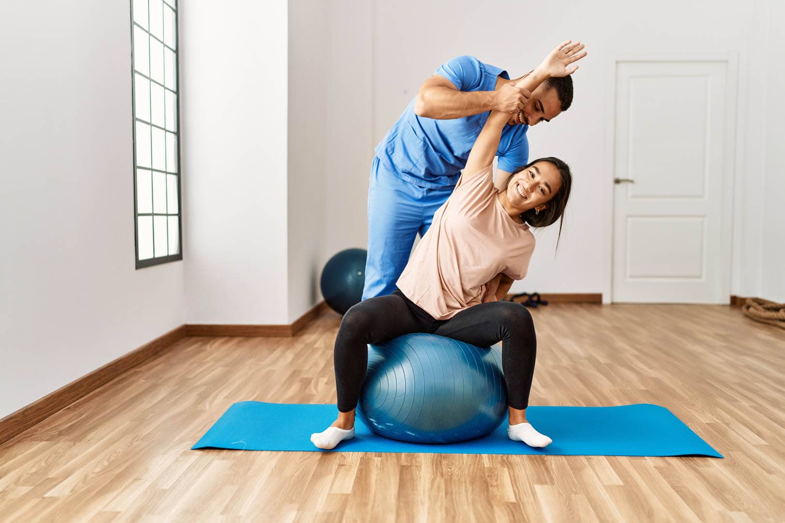 Frau übt Bewegungstherapie auf einem Gymnastikball zu Hause mit Hilfe eines Physiotherapeuten, der ihre Haltung korrigiert.